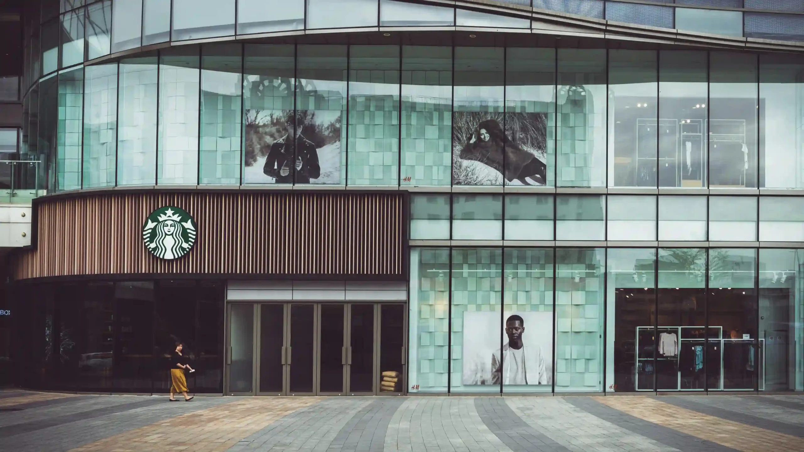开在商场一楼的星巴克店铺外景,玻璃幕墙上有多个广告海报,展示时尚人物,环境现代化。