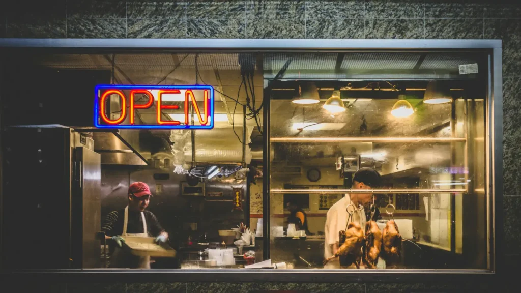 通过餐厅橱窗看到一个霓虹灯‘OPEN’标志。窗内，两名厨房工作人员正在工作；一人正在处理食物，另一人正在照看挂在背景中的烤鸭。