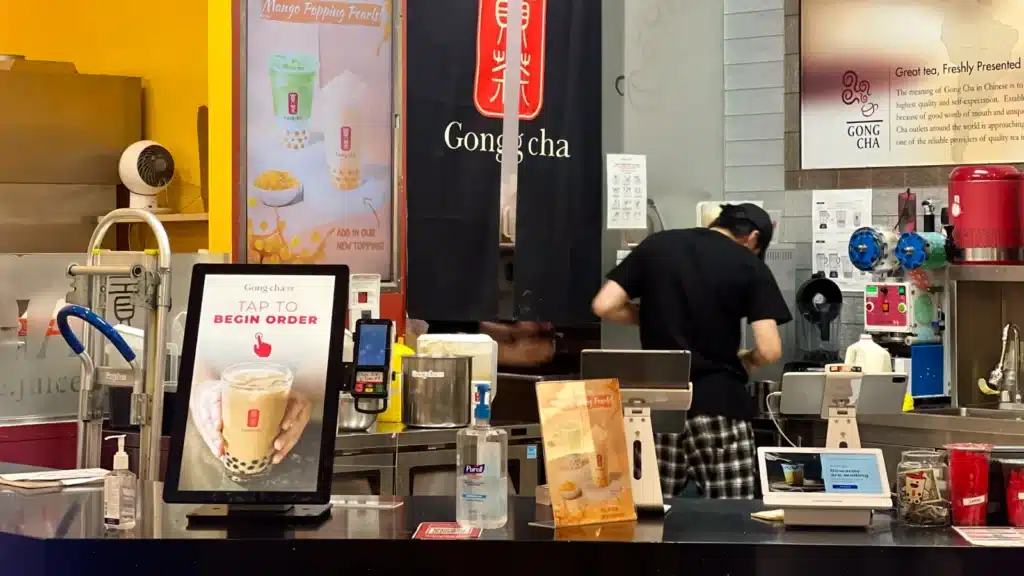 奶茶店的柜台,摆放着一个自助点餐机,一个双屏点餐电脑、消毒洗手液、和奶茶制作设备。柜台后是正在操作的工作人员。背景中有店铺标志和奶茶的宣传海报。