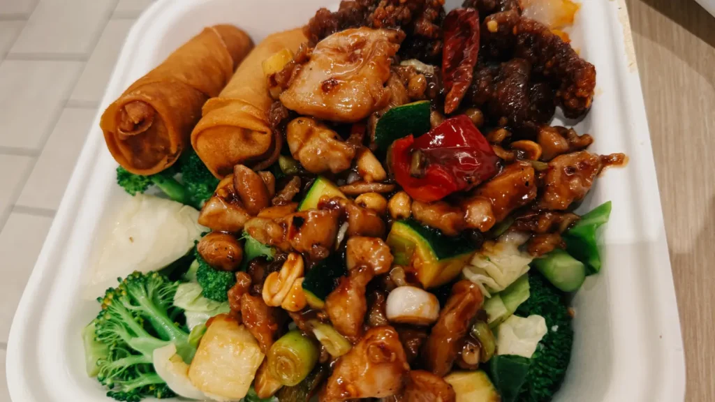 一份色泽鲜艳的中餐盒饭，包括春卷、炒鸡肉、辣椒、花生、蔬菜和西兰花，装在一个外卖盒中。