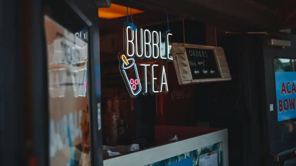 一个奶茶店的前台,挂着一个发光的“Bubble Tea”霓虹灯招牌,旁边还有一个木质的“Orders this way!”指示牌,指引顾客点单的方向。