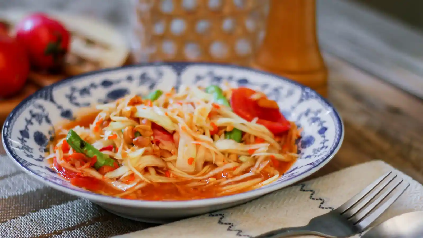 一盘泰国餐馆的面食，面条上铺有切碎的卷心菜、胡萝卜、四季豆和番茄，配上酱汁，色泽鲜艳。餐具整齐地摆放在旁边，整道菜给人一种清新可口的感觉，充满了浓郁的泰式风味。