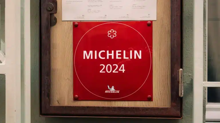 图中红色的标牌上写有“MICHELIN 2024”字样，并且标有一个米其林星级的标志。这块标牌通常悬挂在获得米其林星级餐厅的入口处，象征着该餐厅在2024年度获得了米其林指南的认可。标牌的下方还有米其林轮胎人标志，背景为木质和金属的装饰，整体设计简洁而具有权威性。