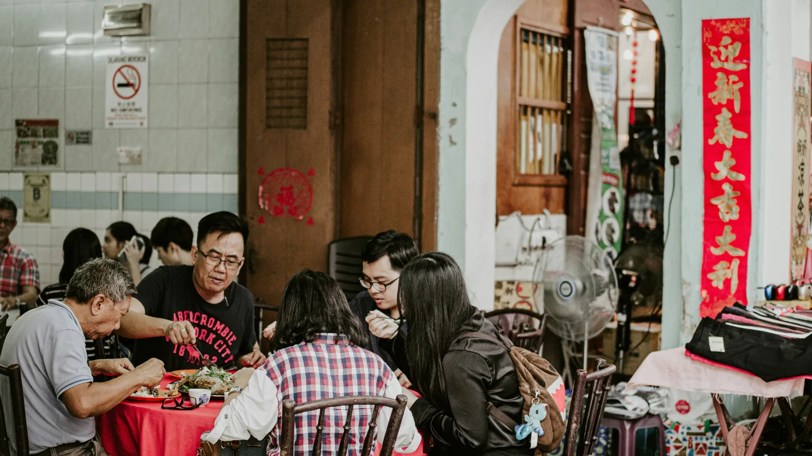 一群人在一家传统华人餐馆里围坐在红色餐桌旁热闹用餐,墙上贴着春节对联,背景是老旧但充满生活气息的餐厅环境,体现了地道的华人饮食文化氛围。