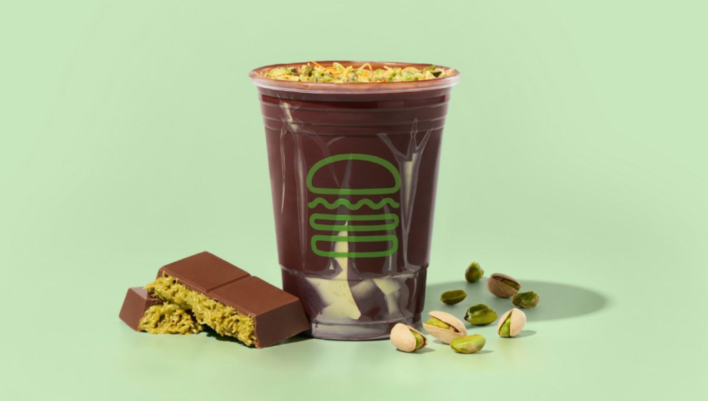 淡绿背景下，一杯迪拜巧克力奶昔顶部撒碎开心果，杯身有品牌图形；旁放切开的巧克力棒，露出开心果与knafeh/kataifi脆丝夹心，并散落开心果仁，强调迪拜巧克力风味。