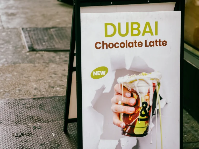 人行道A字板海报，绿色大字“DUBAI”与“Chocolate Latte”，手持一杯淋有巧克力与开心果风味酱的饮品，右侧写有“NEW”，突出迪拜巧克力主题与上新信息。