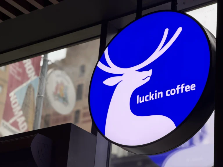 瑞幸咖啡蓝色鹿头标志近景,店内可见曼哈顿街景倒影,象征品牌以科技与效率形象进入美国市场。