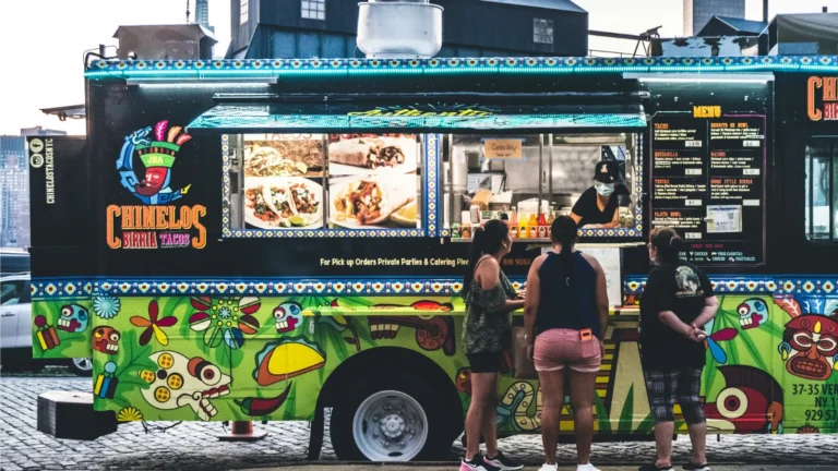 三位顾客在纽约街头的墨西哥风味塔可餐车前点餐,餐车外观色彩鲜艳,展现美国多元街头美食文化。