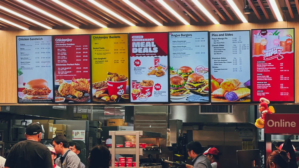 Jollibee 美国门店的点餐柜台现场,顾客排队点餐,背后大型电子菜单展示炸鸡、汉堡、意面与饮品等主要产品。