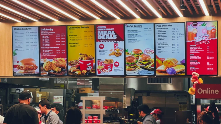 Jollibee 美国门店的点餐柜台现场,顾客排队点餐,背后大型电子菜单展示炸鸡、汉堡、意面与饮品等主要产品。