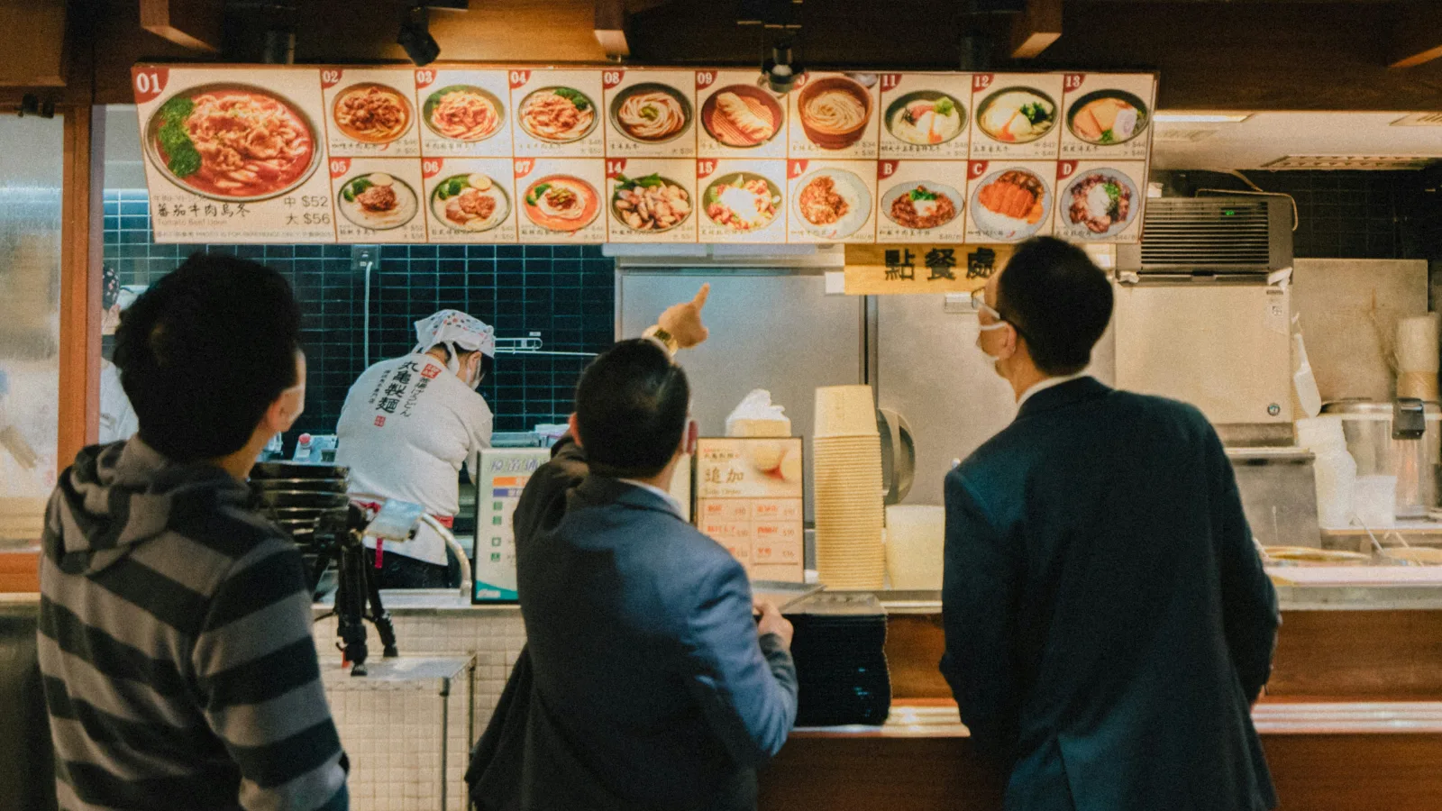 几位顾客站在餐厅点餐柜台前查看头顶菜单板,店员正在后方厨房区域忙碌。
