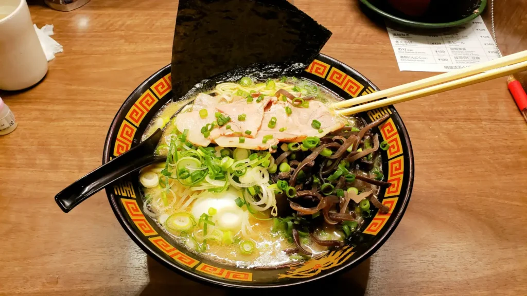A bowl of Japanese ramen on a wooden table, topped with sliced pork, seaweed, scallions, mushrooms, and a soft-boiled egg, showing an in-store dining scene.