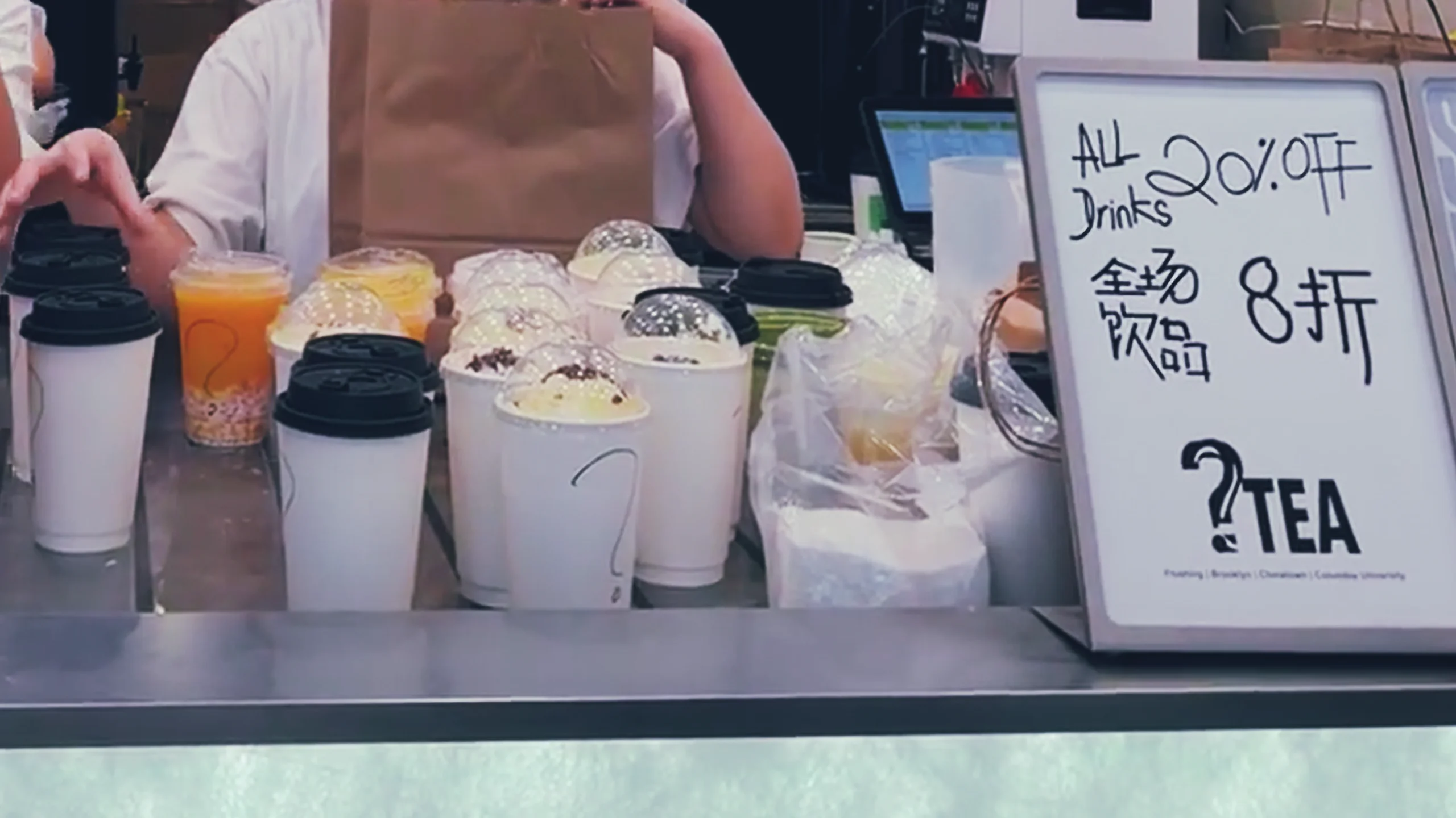 纽约 ?Tea 门店出杯台摆放多杯饮品和全场饮品八折促销牌,杯身使用问号临时标识。